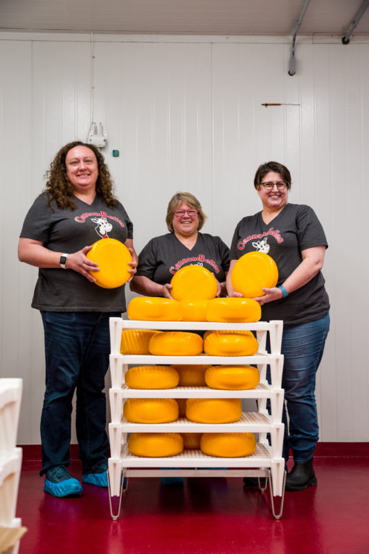 Three women smiling next to a cart full of yellow cheese wheels