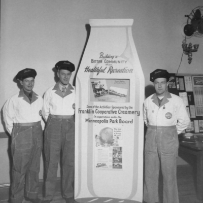 Three milkmen in front of a large milk bottle display