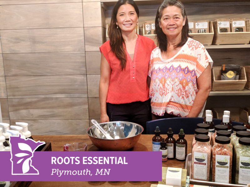 Two people stand next to a table full of wellness products, with text overlay reading "Roots Essential"