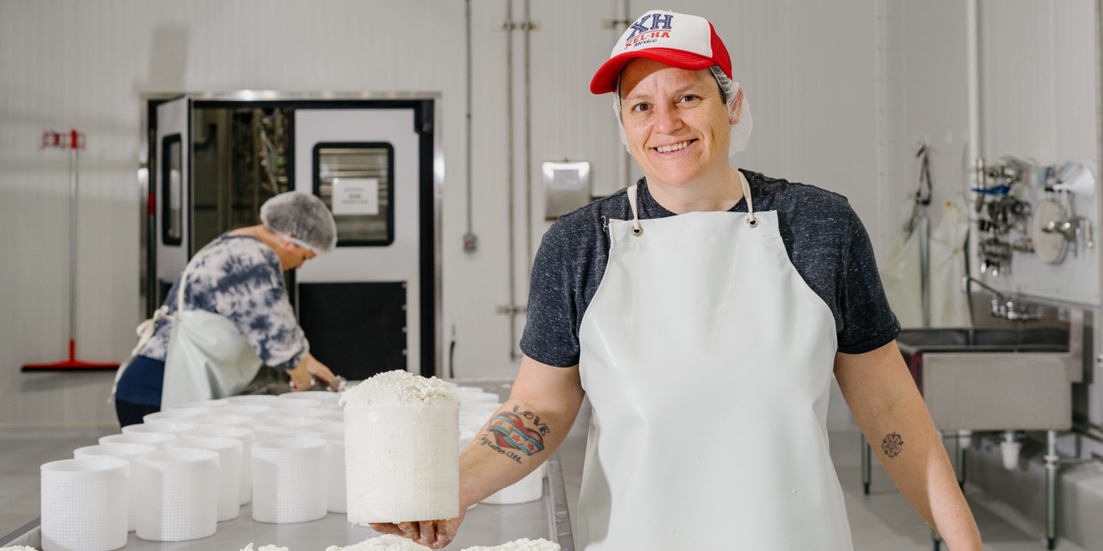 A person in a white apron and a hat holding a tub of goat cheese