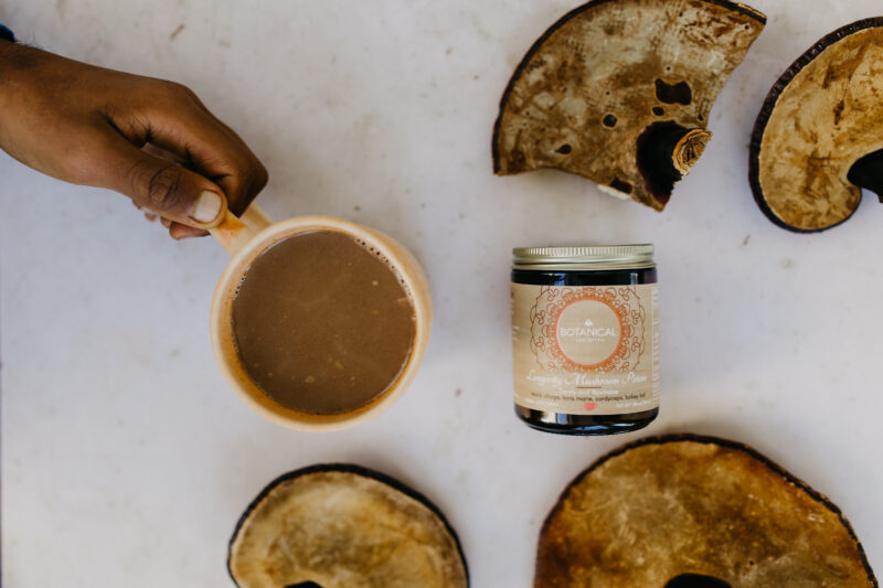 A photo of a Botanical Lucidity elixir with mushroom pieces and a hand holding a mug