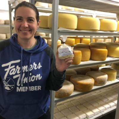 A person smiling and standing in front of shelves full of cheese wheels