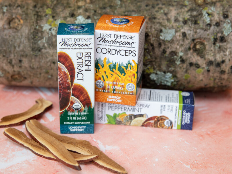 A photo of Host Defense products and mushrooms against a log and a pink background