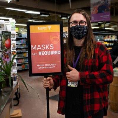 person giving a thumbs-up standing next to a "masks are required" sign