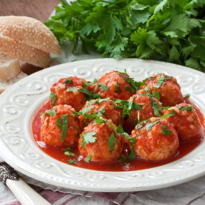 A white plate of turkey meatballs covered in a red sauce with parsley and bread in the background.