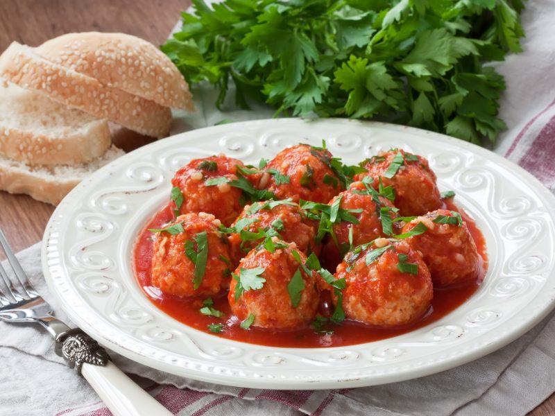 A white plate of turkey meatballs covered in a red sauce with parsley and bread in the background.