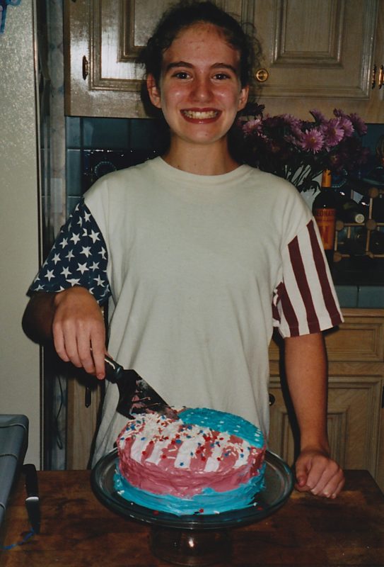 A smiling girl wearing a patriotic shirt and cutting an American flag cake