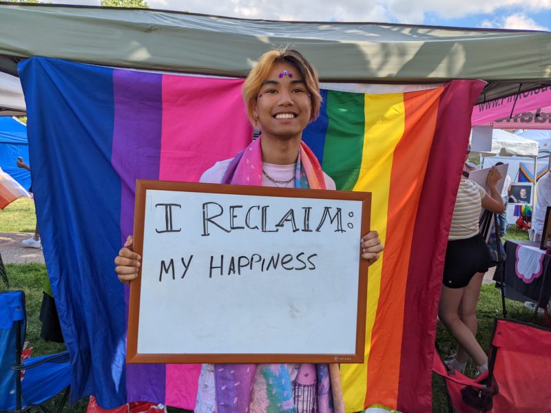 A person in front of a rainbow flag smiling and holding up a sign that reads "I reclaim my happiness"