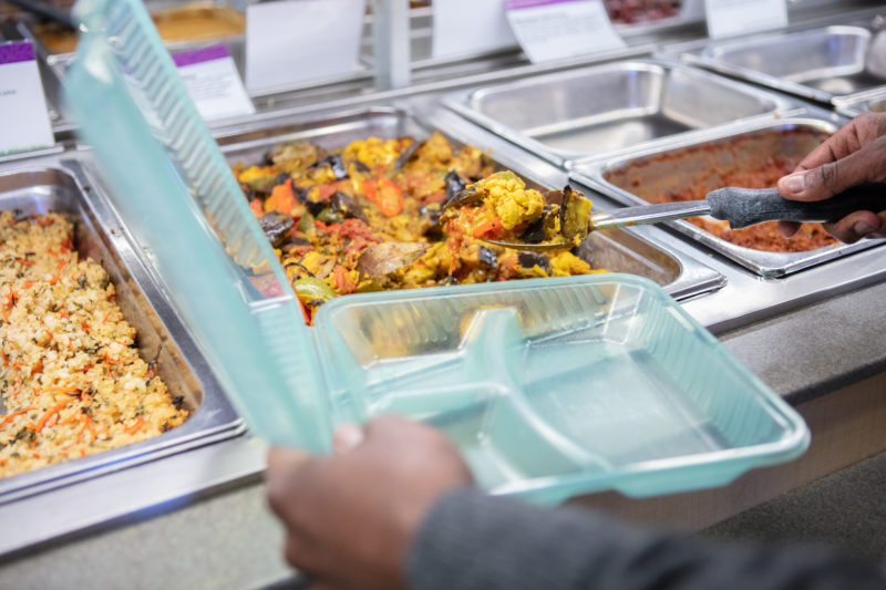A person filling a green reusable hot bar container with food from the Deli