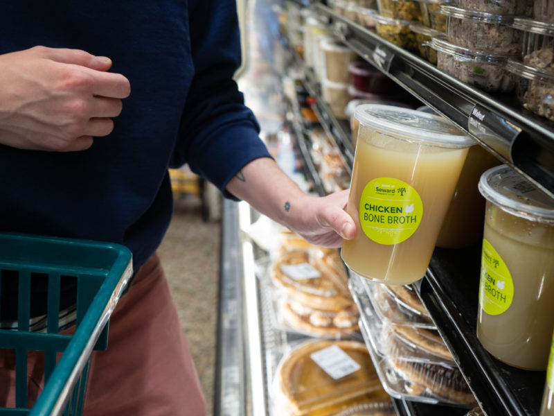A person in a blue sweater takes a container of chicken bone broth off of a refrigerated shelf. The container is labeled with a bright green sticker.
