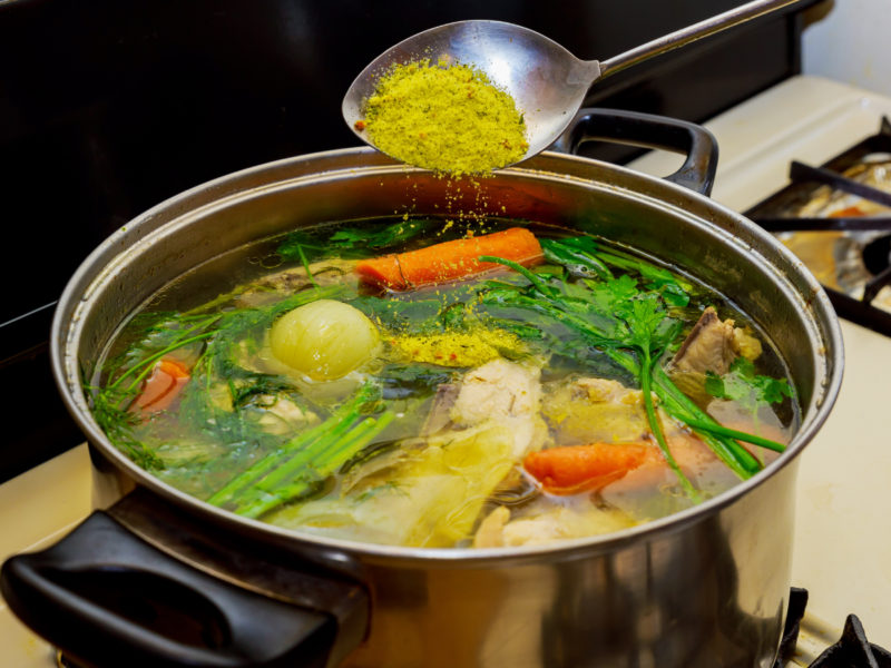A spoon over a colorful pot of boiling green, yellow, and orange vegetables is sprinkling a spice mixture into the broth.