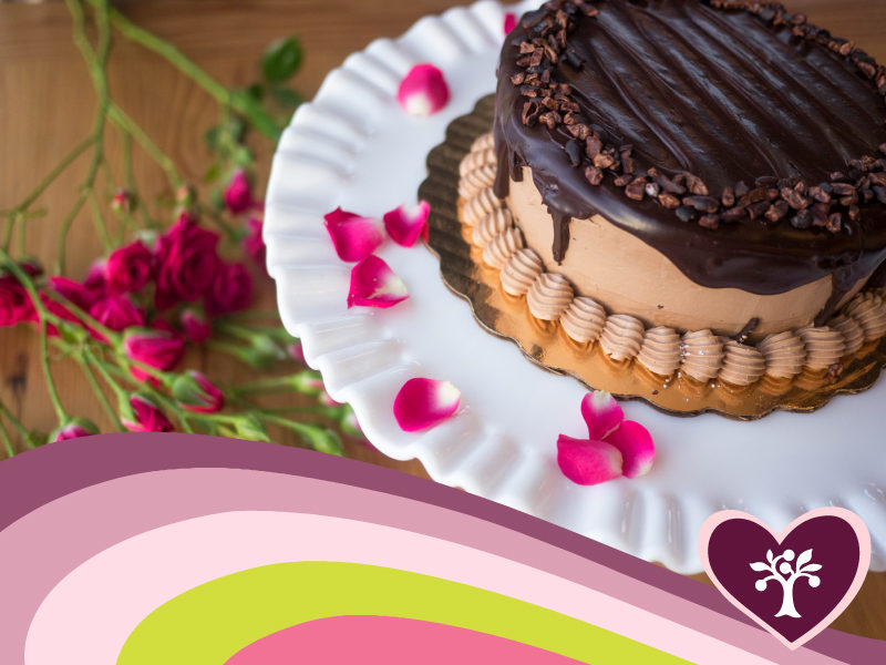 A photo of a chocolate cake on a white stand with rose petals