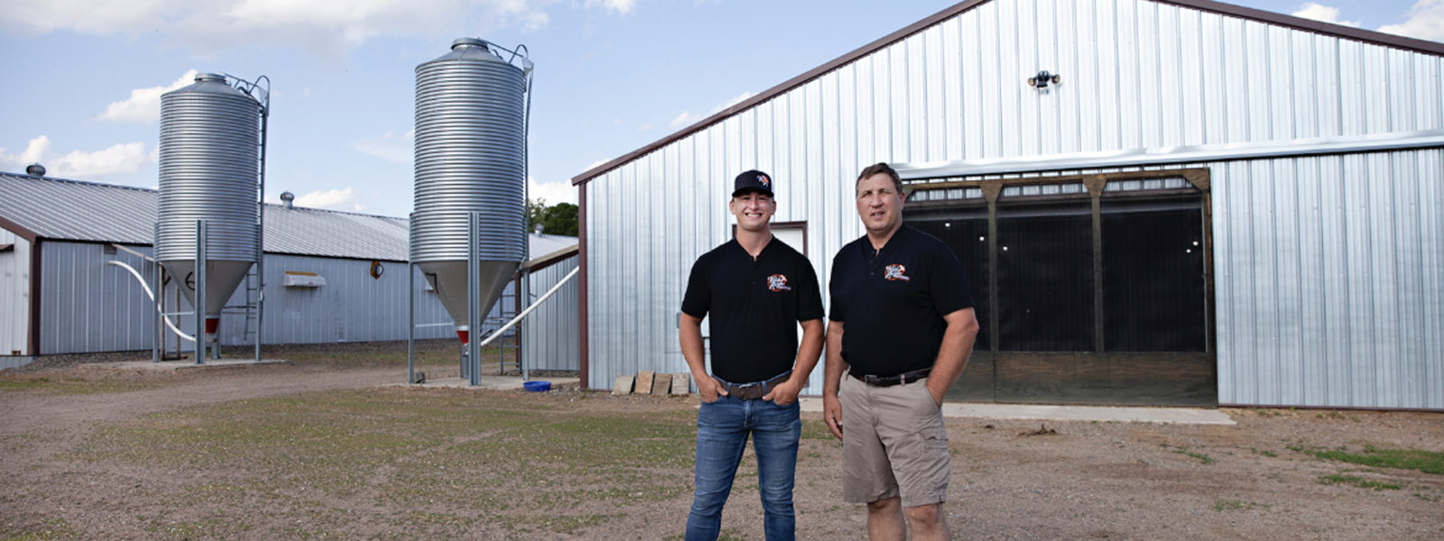 Two poultry farmers wearing black shirts and black hats are standing in front of large silver buildings and equipment on their farm.