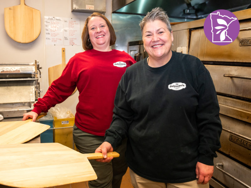 Two women standing in a kitchen smiling and holding pizza paddles