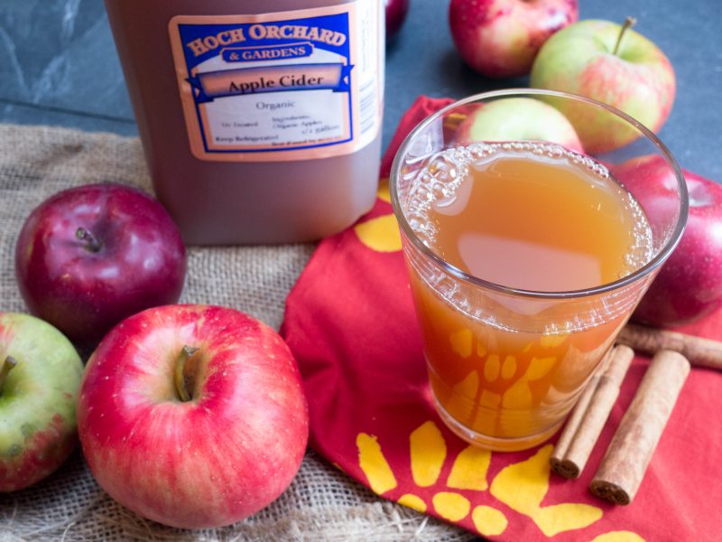 apples next to a glass of apple cider, cinnamon sticks, and a jug of cider