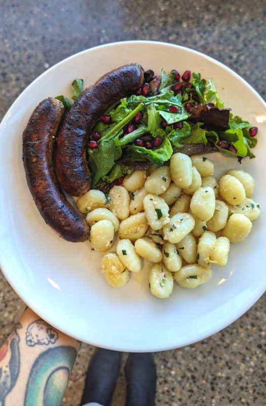 A person holding a plate of sausage with gnocchi and salad