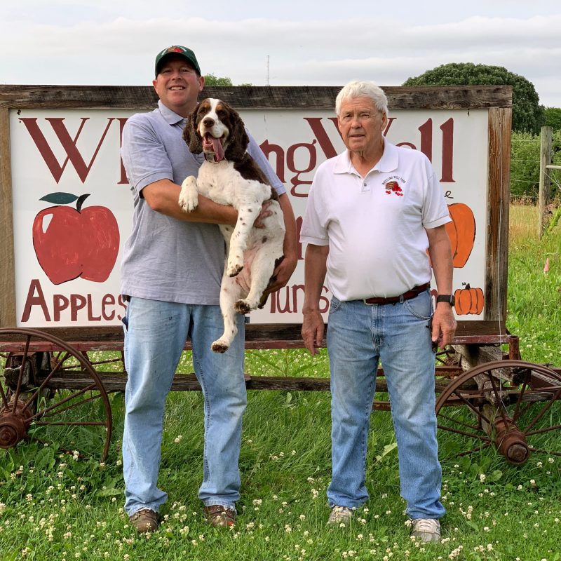 Two farmers holding a dog in front of a sign